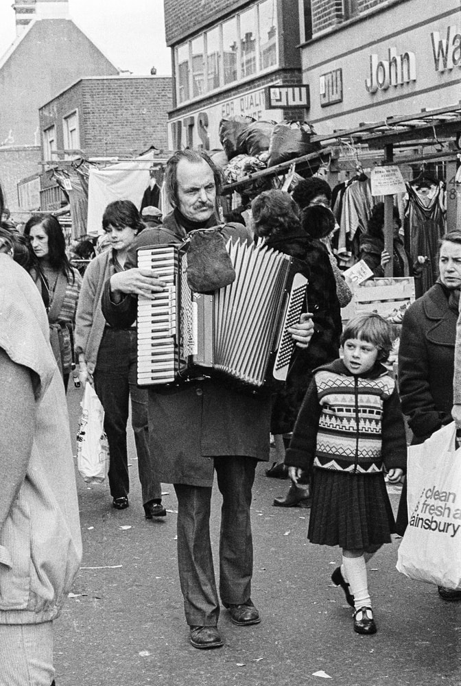 Photo by Brian de Montalk from East End '78 - Roman Road Market, black and white film photos of the East End of London in 1978 at Photospace Gallery Wellington New Zealand September-October 2022, photo of street busker playing accordion in London 1978 black and white film street photography